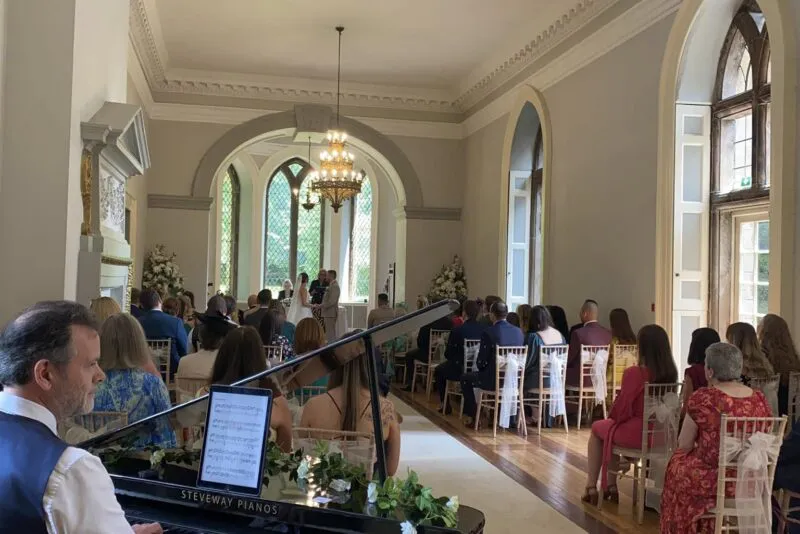 A pianist plays a grand piano in the foreground while wedding guests watch a ceremony unfold at the far end of an elegant, sunlit ballroom.
