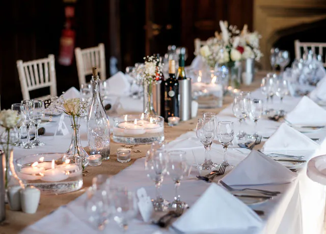 An elegantly styled long banquet table features crisp white linens, folded napkins, wine glasses, floating candles, and delicate bud vases filled with flowers.