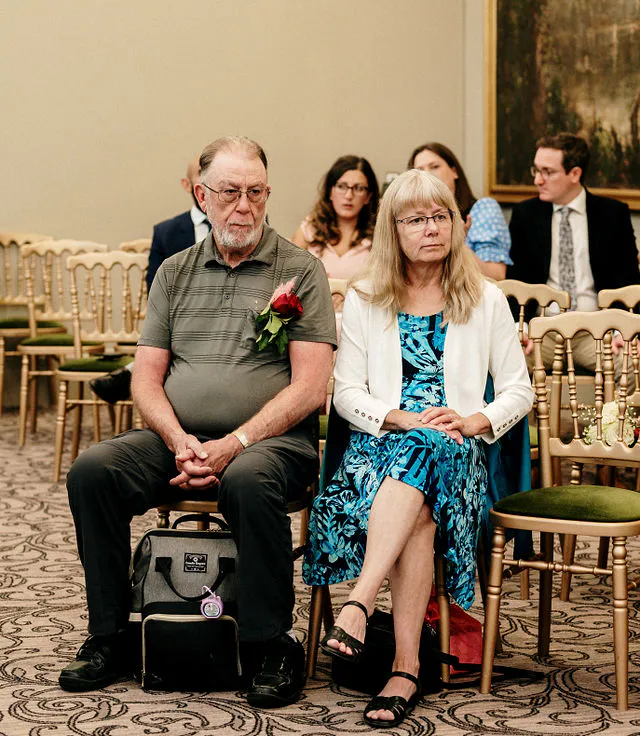 An older couple sitting together as wedding guests, with other attendees visible in the background seated on gold chairs.