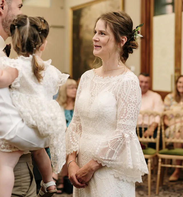 A smiling bride wearing an intricate lace gown with bell sleeves looks tenderly at a young child held by a groom during a heartfelt wedding ceremony.