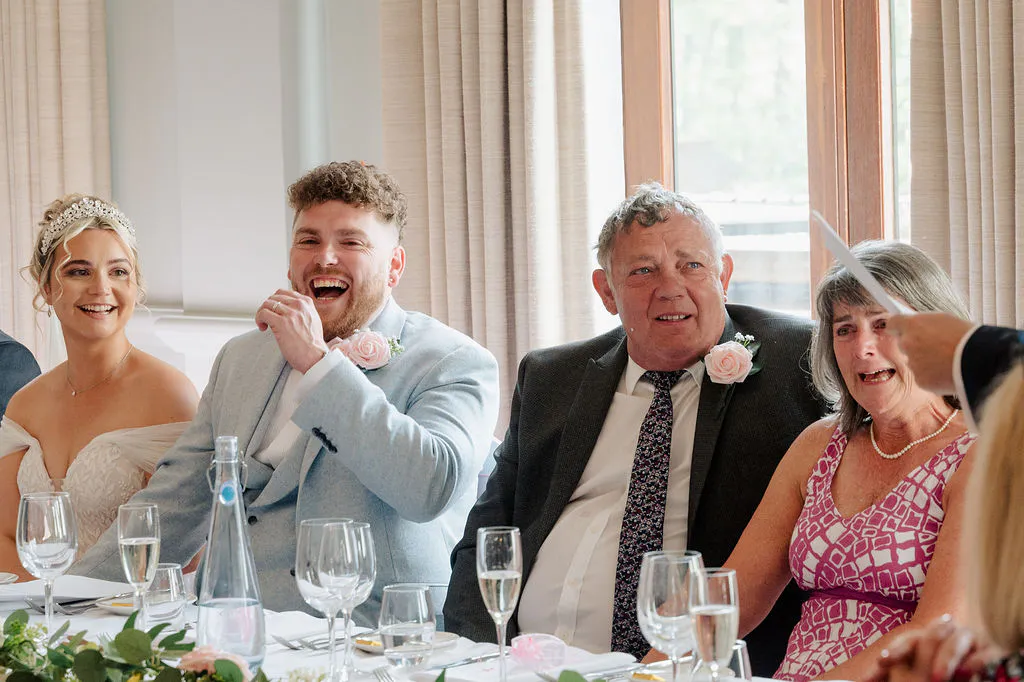A newlywed couple and their parents seated at a wedding reception table, sharing a joyful moment as the groom laughs heartily and the mother of the bride tears up during the speeches.