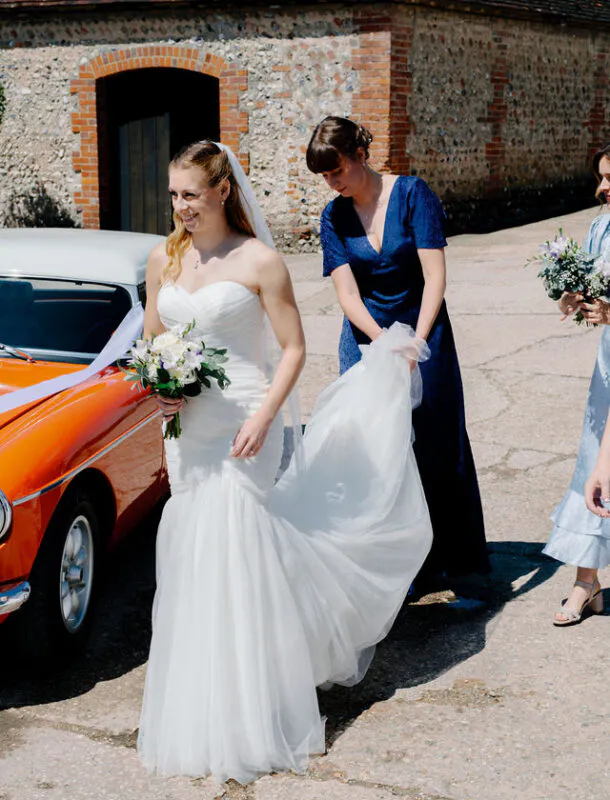 A smiling bride holding a bouquet of white flowers steps out of a classic orange car as a bridesmaid helps adjust her tulle trumpet gown outside a rustic stone barn.