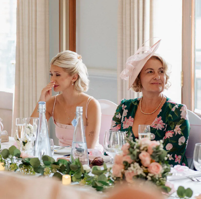 An emotional guest wiping away tears during the wedding speeches at the top table, seated next to an elegant woman wearing a floral dress and a pink fascinator.