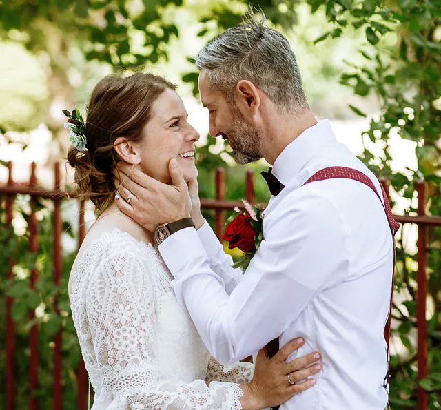 A smiling bride and groom share a tender moment outdoors with dappled sunlight filtering through the trees, wearing a lace wedding dress and a white shirt with red braces.