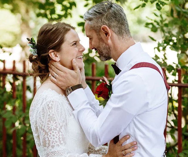 A smiling bride and groom share a tender moment outdoors with dappled sunlight filtering through the trees, wearing a lace wedding dress and a white shirt with red braces.