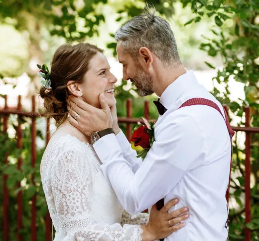 A smiling bride and groom share a tender moment outdoors with dappled sunlight filtering through the trees, wearing a lace wedding dress and a white shirt with red braces.