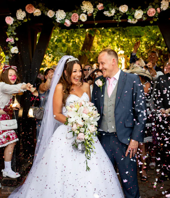 A joyful newlywed couple laughs happily amidst a shower of colorful confetti under a floral archway as they exit their wedding ceremony.