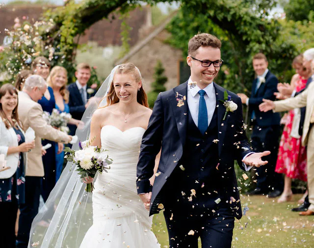 A smiling bride and groom walk hand in hand through a shower of confetti under a stone garden archway, surrounded by cheering guests.