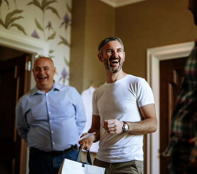 The groom laughing joyfully while holding a gift bag during morning preparations surrounded by family and friends.