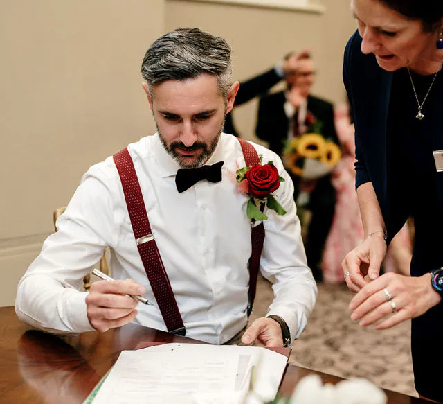 A groom wearing a white shirt, dark braces, a bow tie and a red rose buttonhole signs the marriage register while seated at a wooden table.