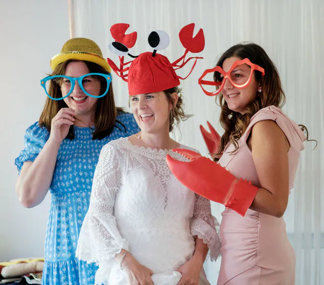 A smiling bride wearing a novelty crab hat poses with her bridesmaids who are wearing funny glasses and holding large toy lobster claws during wedding preparations.