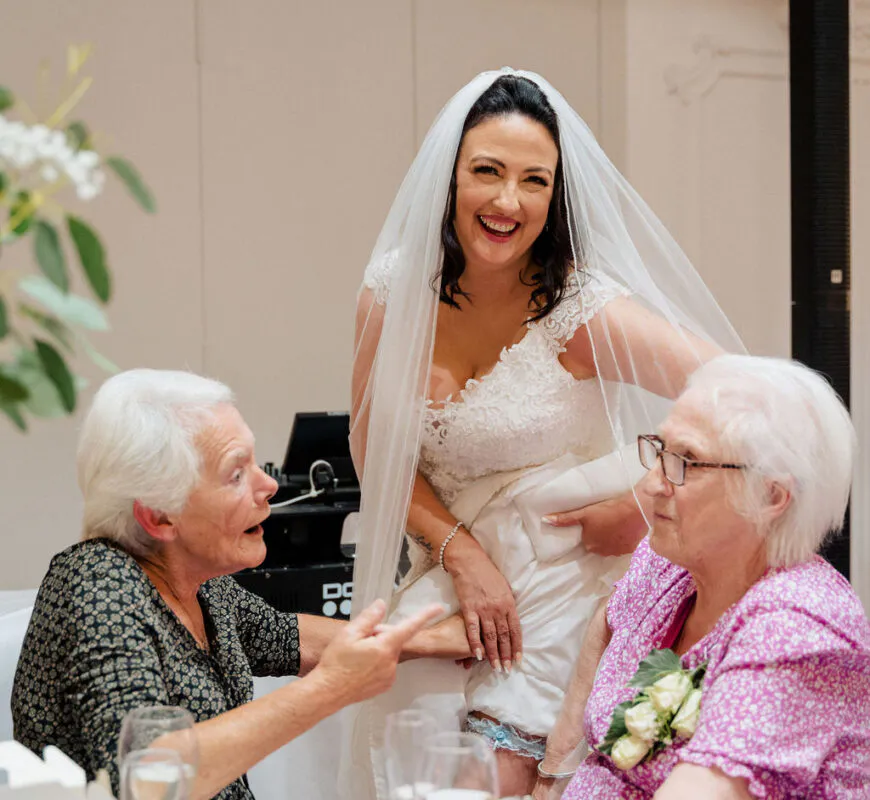 A joyful bride wearing a lace wedding dress and veil laughs happily while standing with elderly guests at a wedding reception.