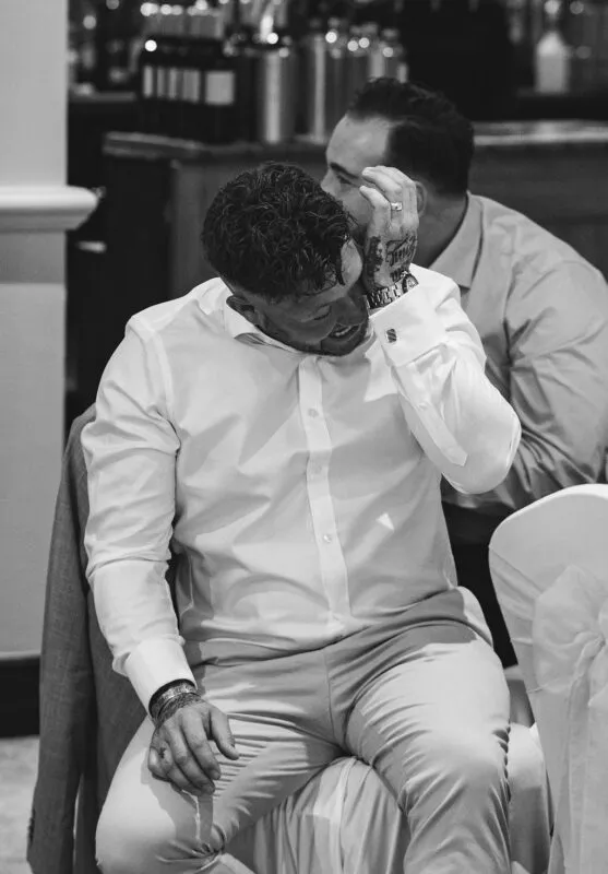 A black and white candid photograph captures a groom seated at the wedding reception, overcome with laughter and wiping a tear during the speeches.