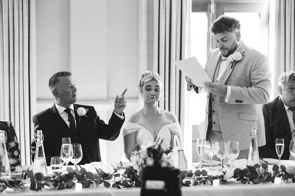 A groom reads from his speech at the top table while a guest reacts with humor and the bride pulls a playful face.
