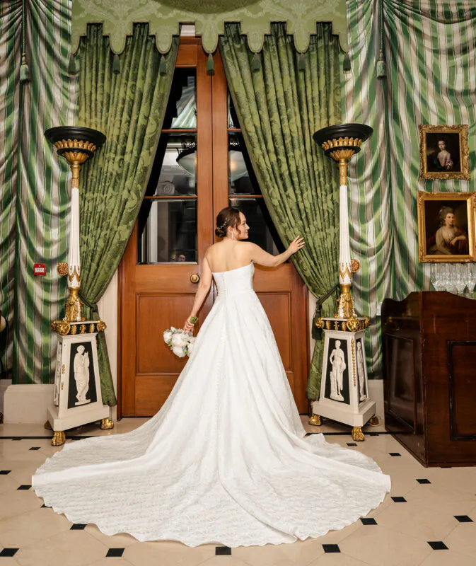 A bride in a strapless A-line wedding dress with a dramatic sweep train stands back-facing in a grand country house interior.