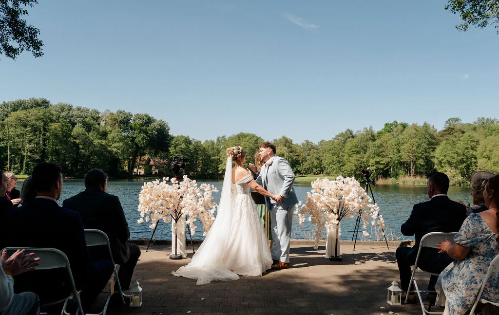 A bride and groom exchange vows during an outdoor lakeside ceremony under a clear blue sky, flanked by white floral displays with guests seated on chairs.