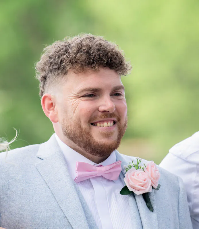 A smiling groom wearing a light grey suit, pink bow tie, and a matching rose buttonhole looks happily towards the ceremony.