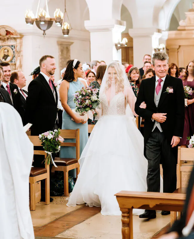 A smiling bride walks down the church aisle holding a bouquet, linked arm in arm with her father who wears a traditional morning suit.