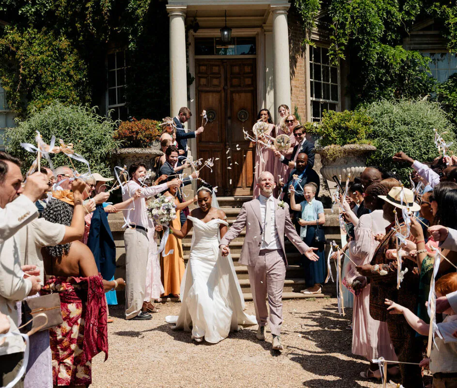 A joyful newlywed couple walks hand in hand down the stone steps of a historic country house while cheering guests wave ribbon wands in celebration.