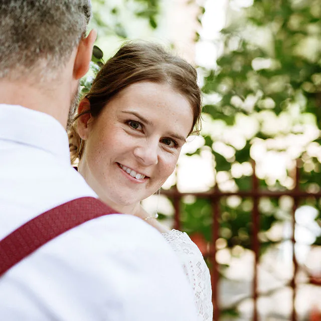 A smiling bride peers over the shoulder of her groom outdoors, wearing a lace dress while he wears a white shirt with red braces.