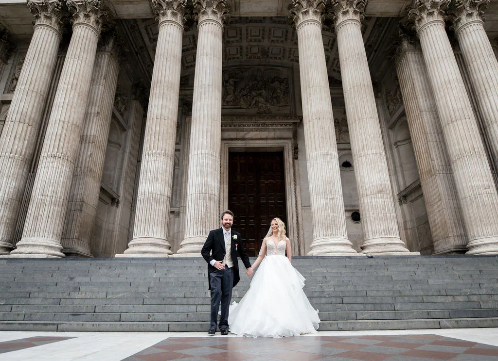 A newlywed couple holds hands while descending the stone steps of a grand cathedral with towering columns.