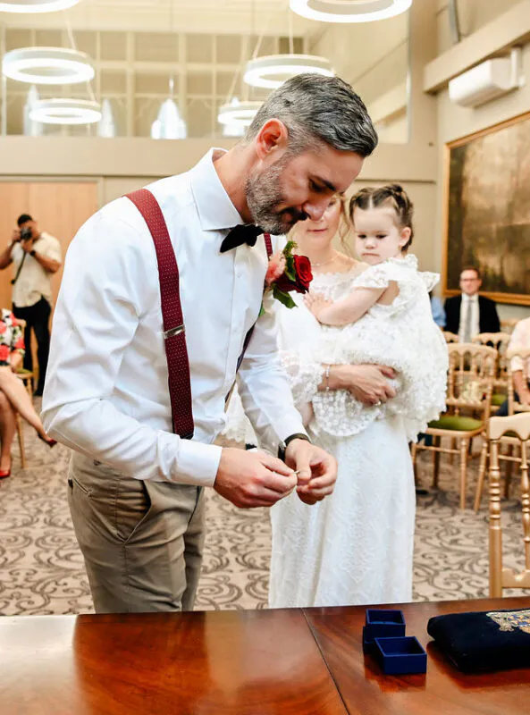 A groom prepares to sign the register during a civil wedding ceremony while the bride holds a young child nearby.