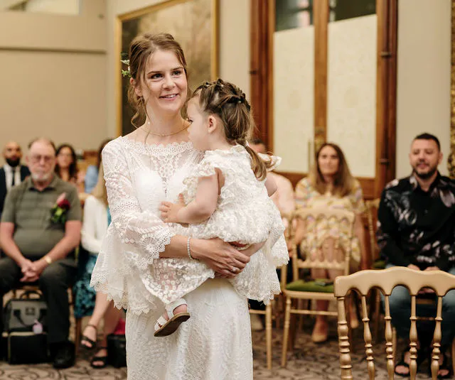 A smiling bride wearing a lace long-sleeved gown holding a young flower girl during a wedding ceremony surrounded by seated guests.