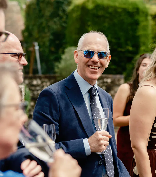 A smiling guest wearing sunglasses and a navy suit holds a glass of champagne during an outdoor wedding drinks reception.