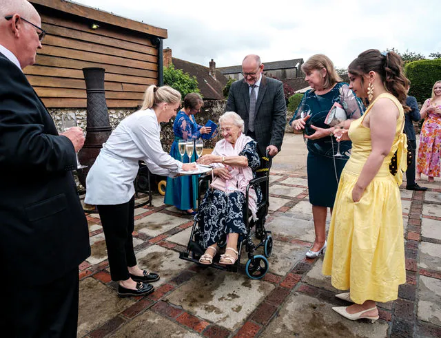 Wedding guests mingle in an outdoor courtyard while a member of the service staff offers champagne to an elderly guest in a wheelchair.
