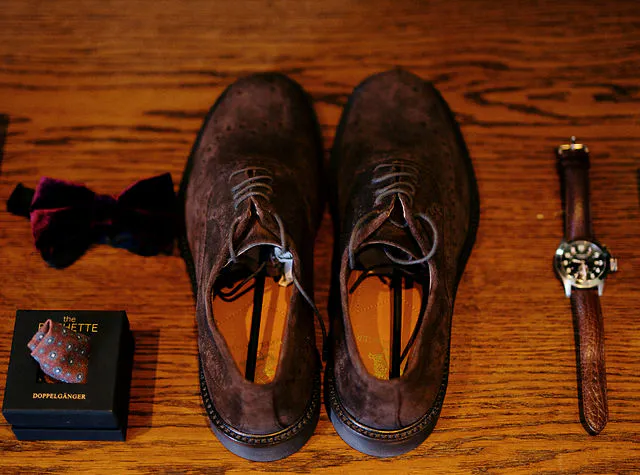 A flat lay of groom accessories featuring brown suede brogues, a dark velvet bow tie, and a leather watch arranged neatly on a wooden table.