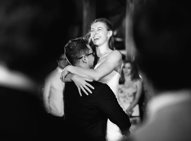 A newlywed couple shares a joyful moment on the dance floor in black and white, with the groom lifting the laughing bride into the air as guests watch on.