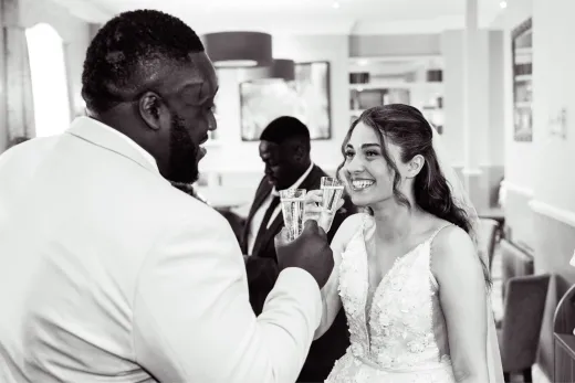 A newlywed couple shares a joyful toast with champagne flutes during their wedding reception, captured in a striking black and white photograph.