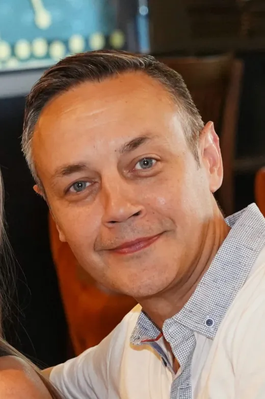 A close-up portrait of a smiling groom with blue eyes wearing a patterned collared shirt during the wedding celebration.