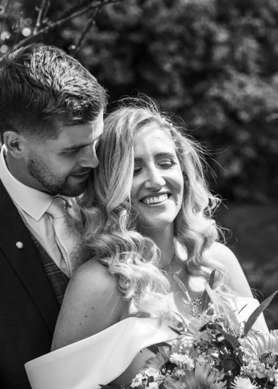 A newlywed couple shares a joyful, intimate moment outdoors with the groom looking lovingly at his smiling bride who holds a bouquet.
