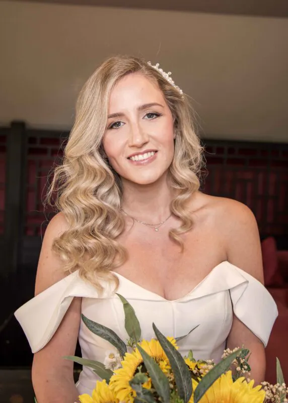 A beautiful blonde bride smiles warmly while holding a vibrant sunflower bouquet, wearing an off-the-shoulder satin gown and delicate pearl hair accessories.