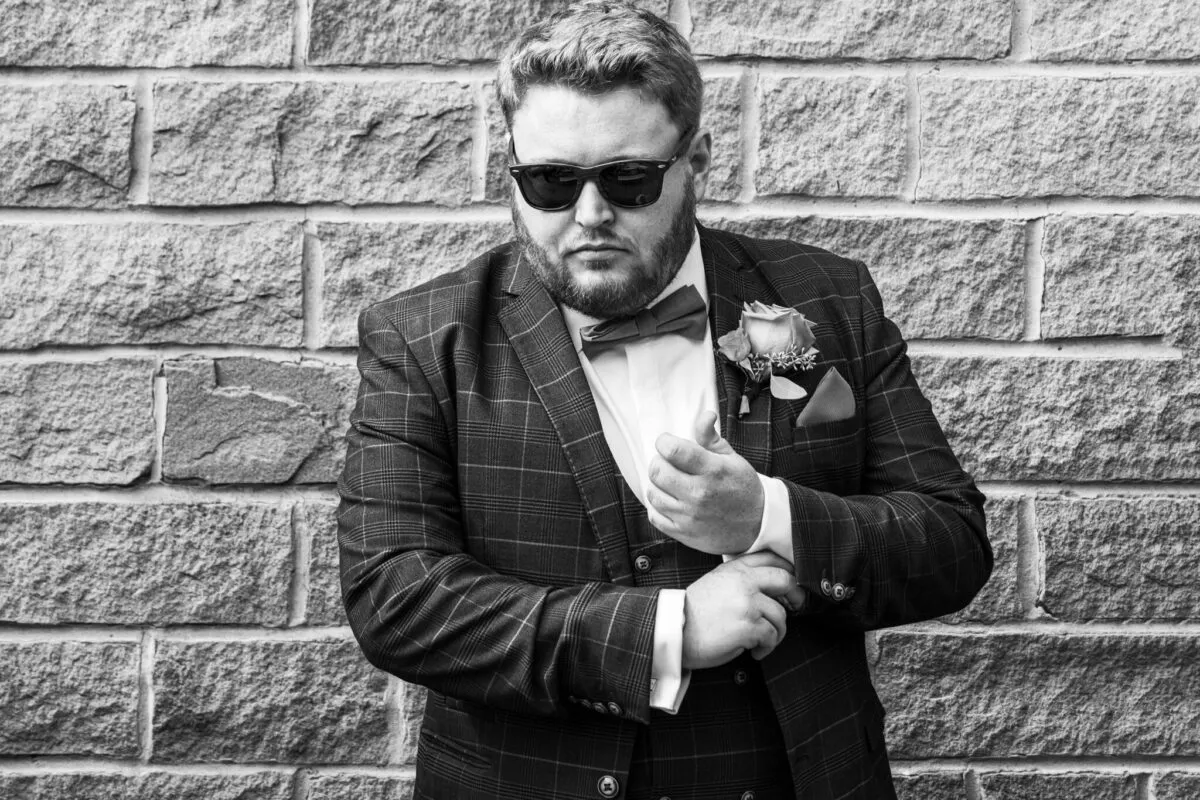 A stylish groom wearing a checked tweed three-piece suit, bow tie, and sunglasses adjusts his cuff against an exposed brick wall in a black and white photograph.