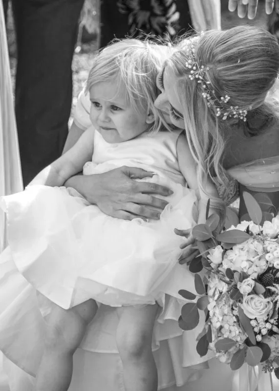 A black and white candid photograph captures a bride holding a young flower girl who is wearing a sweet white dress, with a floral bouquet visible in the foreground.
