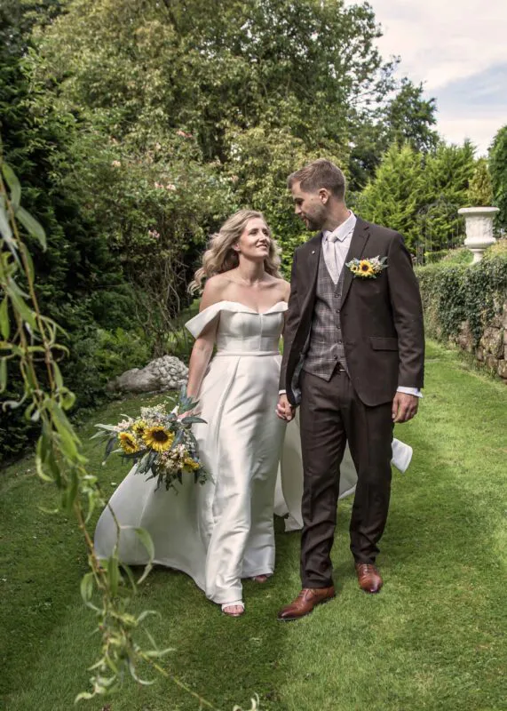 A bride in an off-the-shoulder satin gown holding a sunflower bouquet and a groom in a brown suit walk hand in hand through a lush garden.