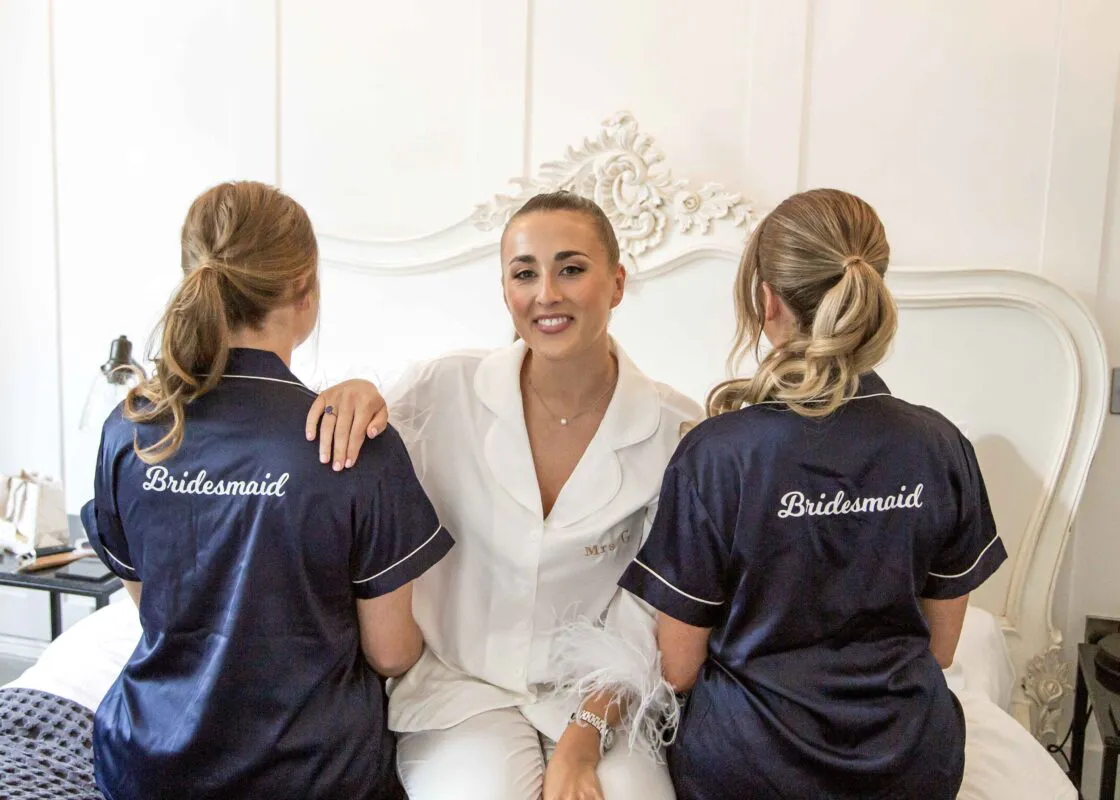 A smiling bride in a white pyjama set with feathered cuffs sits on a bed flanked by two bridesmaids wearing matching navy blue pyjama tops with bridesmaid embroidery.