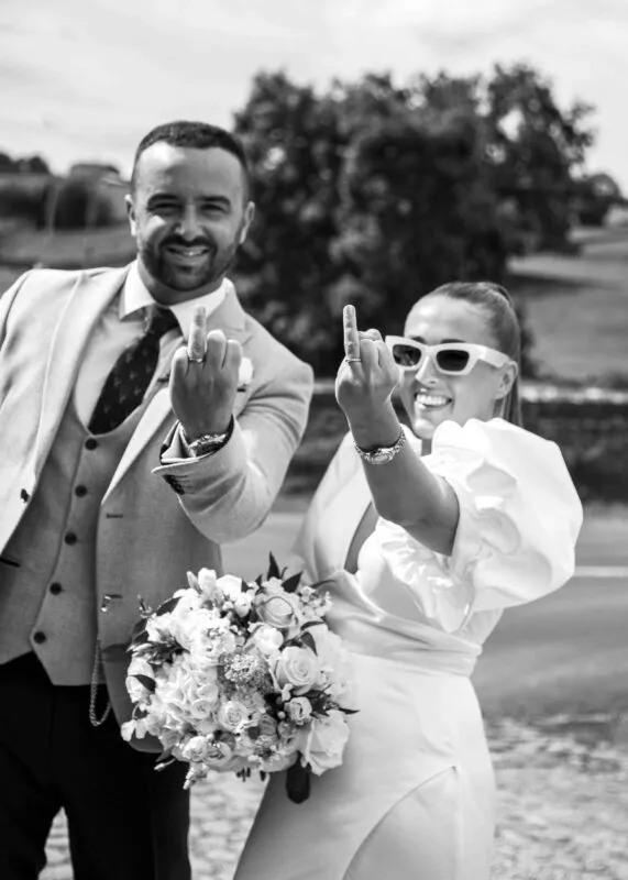 A newlywed couple laughing outdoors in black and white, holding up their middle fingers to showcase their wedding bands while the bride holds a floral bouquet.