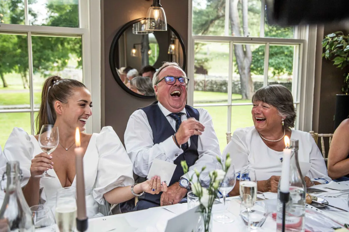 Family members and guests share a moment of pure laughter and joy during the wedding speeches at a candlelit top table.