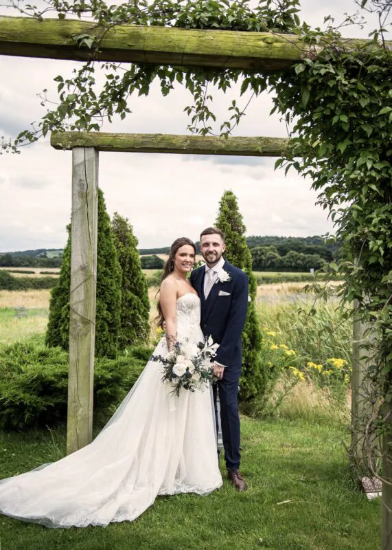 A newlywed couple poses under a wooden garden archway with a lush rural backdrop during their summer wedding portraits.