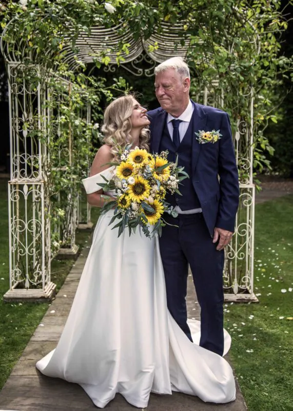 A smiling bride holding a vibrant sunflower bouquet looks up affectionately at her father in a navy suit beneath an ornate garden pergola.