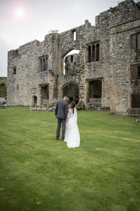 A newlywed couple holding hands stand on a green lawn with their backs to the camera, framed by an ancient stone castle ruin under a soft sky.