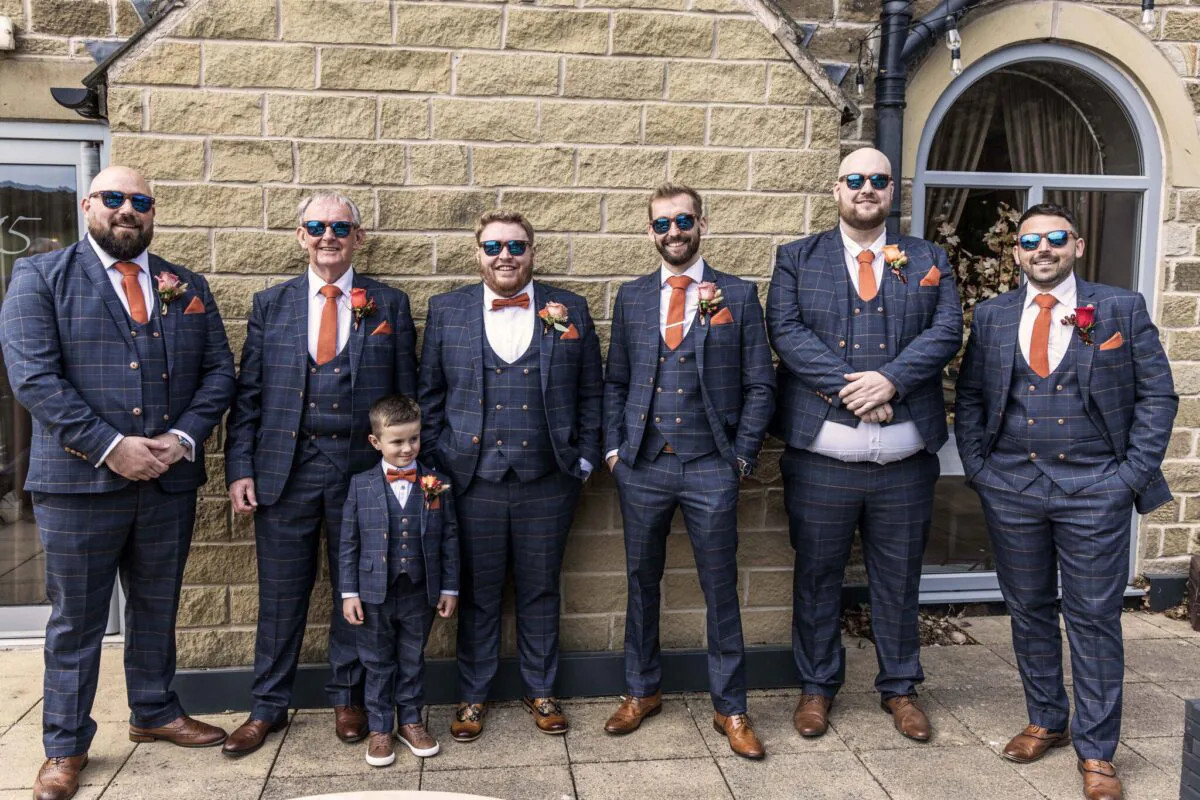 A group of groomsmen and a page boy in matching checked three-piece suits with burnt orange ties and sunglasses pose outside a stone barn.