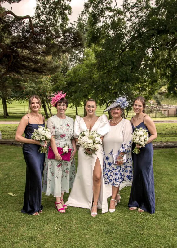 A smiling bride poses outdoors with her mother, older relatives, and bridesmaids wearing navy blue satin dresses and holding white floral bouquets.