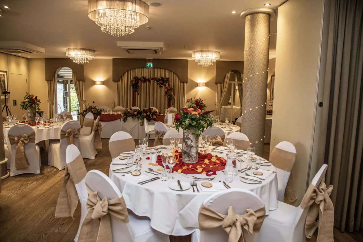 An elegant hotel wedding reception room featuring round tables adorned with white linens, burlap chair sashes, and vibrant autumnal floral centrepieces.