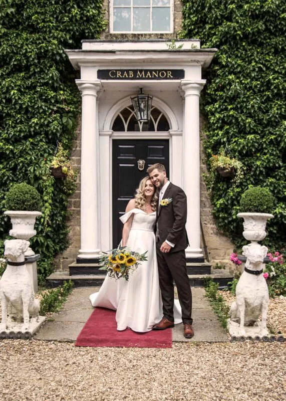 A smiling bride and groom pose outside the entrance of Crab Manor, framed by an ivy-clad stone facade and ornate white statues.