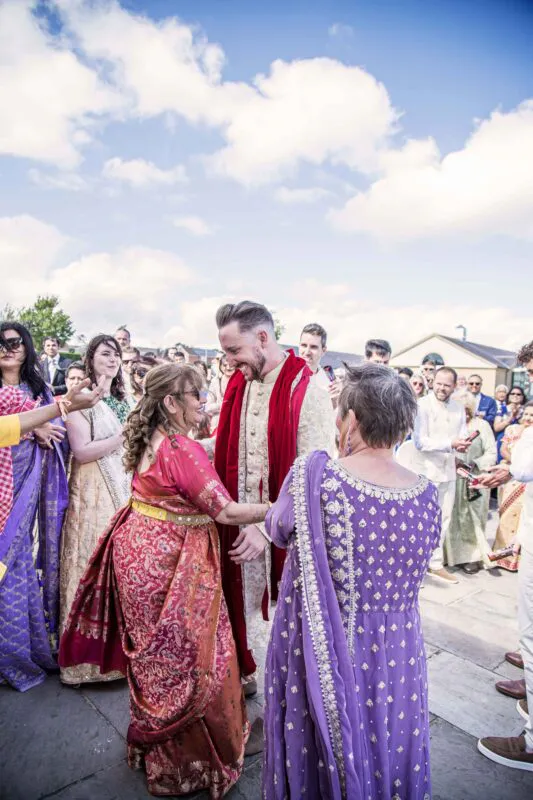 A groom wearing a traditional sherwani with a red stole is greeted joyfully by family members in vibrant traditional attire during an outdoor wedding ceremony.