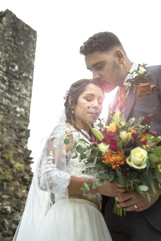 A groom gently kisses the bride's forehead outdoors near stone castle ruins while she holds a colourful autumnal bouquet.